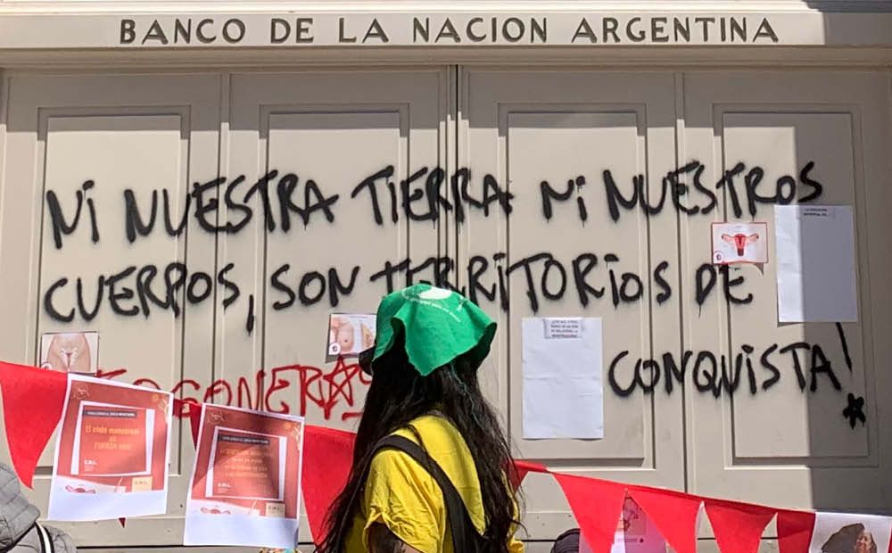 Person with the green bandana of the 2003 National Campaign for the Right to Legal, Safe, and Free Abortion. The graffiti on the wall reads Ni nuestra tierra ni nuestros cuerpos, son territorios de conquista! (Neither our land nor our bodies are territories of conquest!). October 9, 2022, San Luis.( Photograph by Maya Ober)
