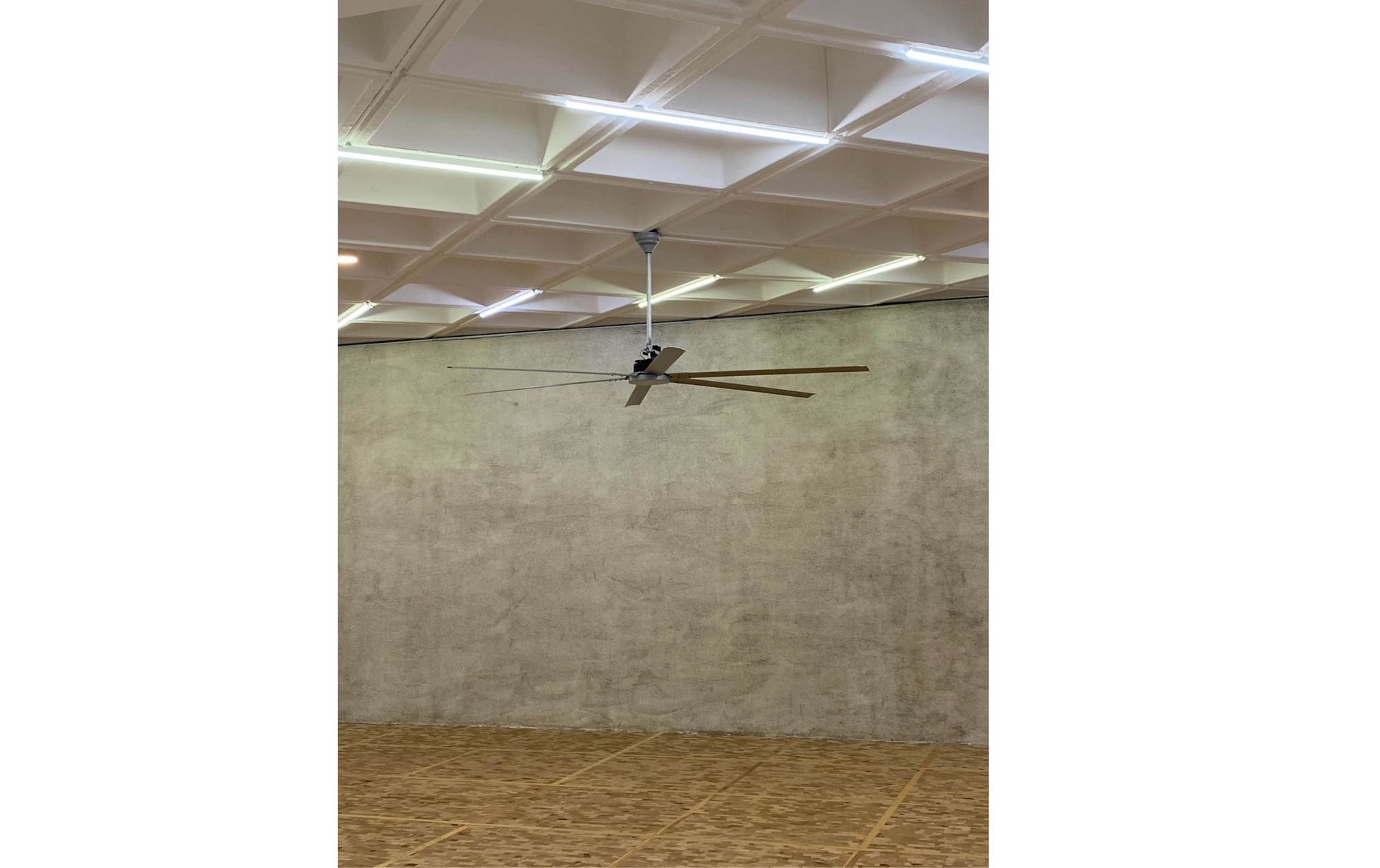 In a stark exhibition space with wooden floors and concrete walls and ceiling, a ceiling fan drops down in the center of the image, seemingly still. 